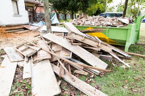 A pile of discarded wooden planks and broken timber, some with chipped edges and weathered surfaces, scattered on a grassy outdoor area in front of a green skip overflowing with mixed construction debris, including bricks and rubble. The scene is set on a residential property with trees and a partially visible house structure in the background, indicating a rubbish removal activity in progress. The wooden materials vary in length and thickness, with some pieces leaning on each other or lying flat among the grass, suggesting recent clearance work by Commercial Waste Kentish Town for a property in the N19 postcode area. Natural daylight illuminates the scene, casting soft shadows on the ground, with the environment reflecting typical waste collection and disposal context familiar to local rubbish removal services.
