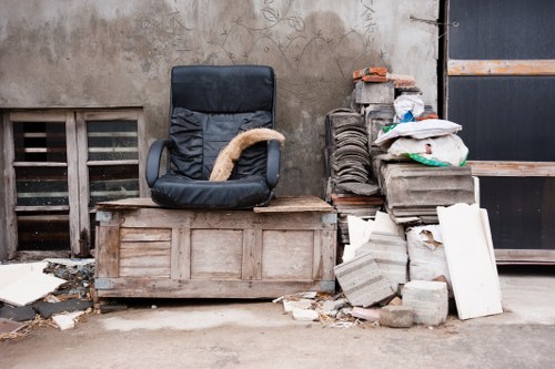 The image shows a makeshift outdoor rubbish collection area situated against a weathered, grey concrete wall. In the foreground, there is a black office chair with visible wear, placed on a wooden pallet that serves as a makeshift platform. To the right of the chair, a pile of discarded construction debris and waste materials is stacked, including concrete blocks, broken tiles, and pieces of white plasterboard, some leaning against a small wall of stacked bricks. The surface below the objects is a rough, concrete ground with scattered small debris and dirt. In the background, there is a grated vent near the wall and a large dark metal door or shutter with a wooden plank running horizontally across it, typical of a maintenance or back entrance area. The scene suggests a location where Commercial Waste Kentish Town might collect or temporarily store waste materials awaiting removal, highlighting typical rubbish in a trades or construction context within the vicinity of Kentish Town.