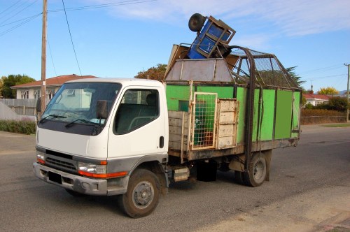 A small white commercial truck parked on a paved residential street with a green cargo area loaded with various discarded furniture items. The truck's cargo space contains a faded wooden chair, a blue plastic storage bin, and a large metal frame, all piled without particular order. The vehicle is positioned near a curb with modest front gardens and residential houses visible in the background, under a clear sky with some light clouds. The truck appears used, with visible dirt and wear on the tires and body, and is part of a rubbish removal service likely involved in clearing household or garden waste from the area. The environment suggests a suburban setting, and the image highlights the process of waste collection and disposal undertaken by Commercial Waste Kentish Town for local clients.