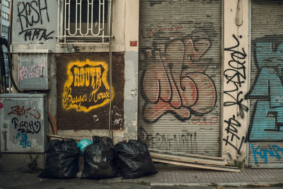 A large pile of black plastic rubbish bags is stacked against a building wall, with some bags partially torn open revealing waste inside. The bags vary in size and are tightly tied at the top, with a